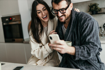 Happy couple laughing and using smartphone in the kitchen wearing bathrobes