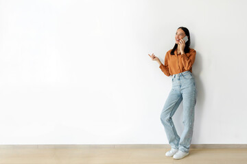 Smiling lady having conversation on cellphone, standing against white wall, talking with client or friend, full length, free space