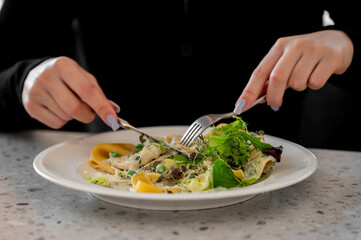 A person eating pasta with leafy greens, peas, and creamy sauce using fork and knife. The close-up captures a dining moment with a focus on fresh and healthy food