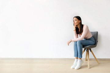 Annoyed sad young lady sitting on chair against white wall, banner with free space. Woman feeling...