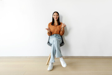 Obraz premium Confident lady sitting on chair, gesturing and talking at camera, isolated on white wall background, having interview, blogger streaming, full length