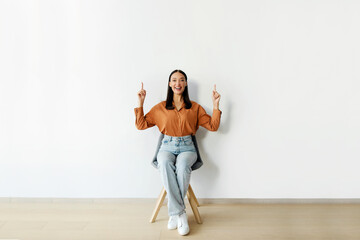 Positive lady sitting in chair over white wall, pointing up at copy space for text or...