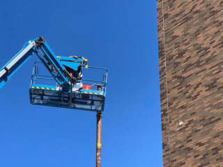 Partial view of a mobile crane high up in the air, situated next to the brick wall of a new building. The platform is unloading a metal beam. Clear blue skies in the background of view.