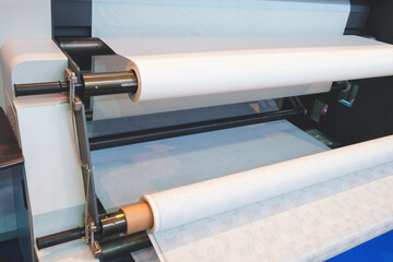 A roll of paper is being fed through a industrial printer machine. The roll is white and the...