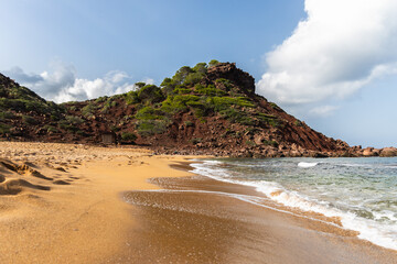 Empty beach Cala del Pilar in Menorca, red mountain and orange sand with crystal clear sea