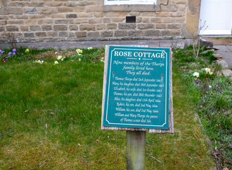 Eham, Chesterfield, United Kingdom - 21 Mar 2025 - A green sign with a poem on it is placed in a grassy area. Passing of members from the black death. The sign is surrounded by flowers, which adds to 
