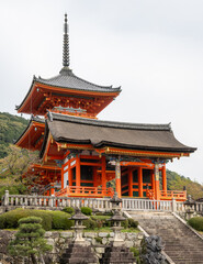 Kiyomizudera's Pagoda Sanjunodo and Nishimon Gate in Kyoto, Japan. A three-tiered vermilion pagoda with gray tiled roofs and ornate spire stands elevated on stone foundations amid manicured gardens.