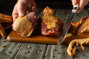 A person expertly slices a piece of poultry on a wooden cutting board surrounded by various chicken parts and spices in a cozy kitchen during the morning hours