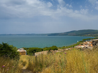 Fototapeta premium Trevignano Romano, Lazio, Italy - July 7, 2024: Lake Bracciano north shore from Orsinis Fortress. Modern residential buildings down the hill. Forested shoreline on other side, under blue cloudscape