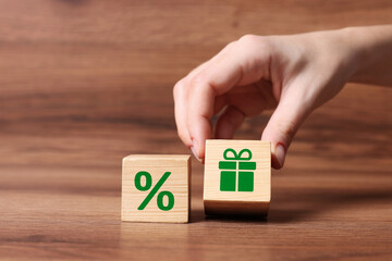 Bonus. Woman taking cube with gift box image near other one with percent sign on wooden table, closeup