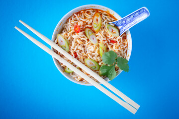 Overhead shot of homemade ramen soup, flavored with chili peppers, cilantro, and scallions, styled with chopsticks on a bold blue surface. Perfect stock image for food marketing and menu layouts.