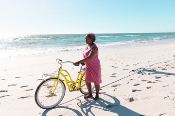 Naklejka premium Elderly woman in pink dress walking yellow bicycle along sunny beach