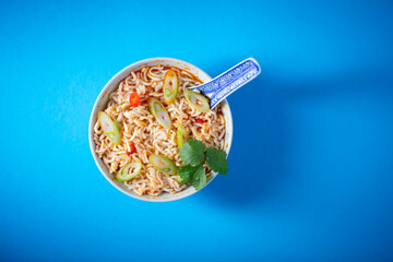 Instant noodle bowl topped with chili, cilantro and onion on blue background. Top shot, nobody.
