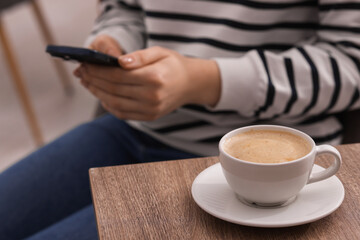 Woman using smartphone during coffee break at wooden table indoors, closeup