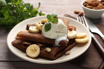 Delicious chocolate crepes with banana, nuts, mint and scoop of ice cream on wooden table, closeup