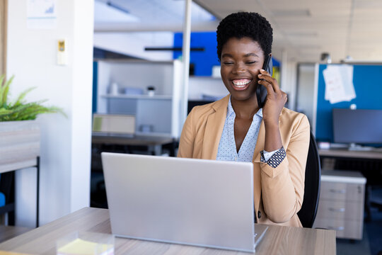 African American woman using smartphone and laptop, smiling at office desk