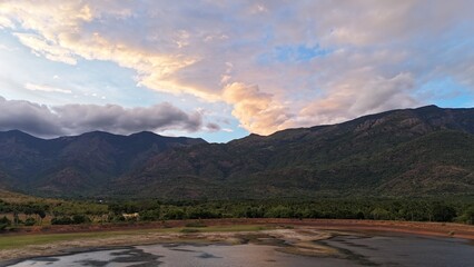 A serene reservoir at sunset with a vibrant sky, scattered clouds, and calm water. Lush greenery and hills enhance the scenic beauty, creating a peaceful natural view.
