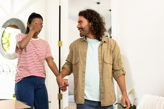 Young diverse couple holding hands, woman playfully covering eyes, entering new home - Powered by Adobe