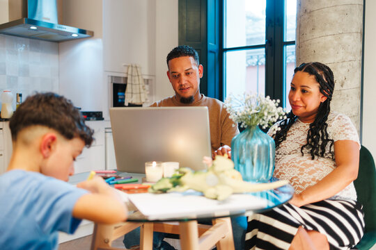 Latin family using laptop in the kitchen while pregnant mom relaxing and child drawing