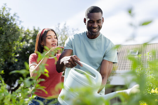 Diverse couple enjoying gardening together, watering plants and smiling in backyard - Powered by Adobe