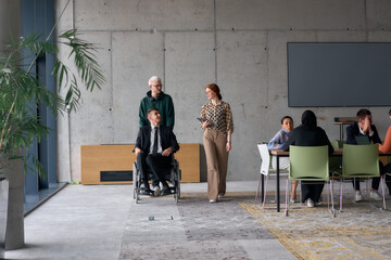 A diverse team of business colleagues, including a director in a wheelchair, engages in a collaborative discussion as they move together through a modern office space