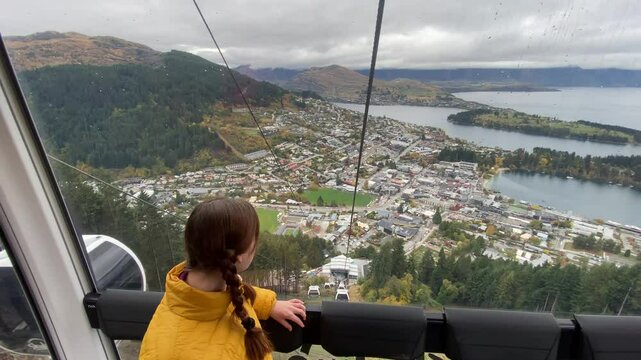  happy girl in yellow jacket sits inside a cable car / gondola going up and enjoys a stunning panoramic view of Queenstown and Lake Wakatipu. Skyline Queenstown, Otago, New Zealand, South Island