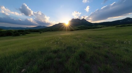 Serene mountain meadow at sunset.  Vast grassy plain stretches towards a mountain range.  Sunlight bursts through clouds, illuminating the peaks.  Delicate wildflowers dot the landscape