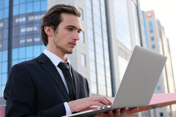 Confident businessman near a business center with a computer. A young businessman in a suit is looking at a laptop. A young businessman near a modern tall glass building with a camputer.