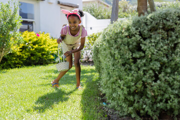 Easter egg hunt, african american girl wearing bunny ears collecting eggs in garden, smiling