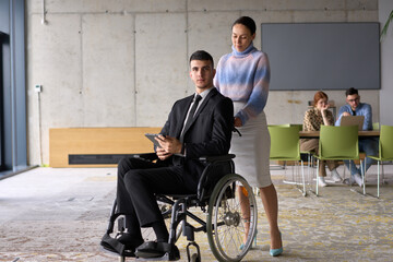 In a professional office setting, a businesswoman helping her director, who is sitting in a wheelchair and using a tablet, while their colleagues collaborate in the background.