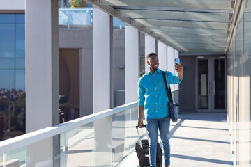 Obraz premium African American man walking with suitcase, taking selfie at modern office building, copy space