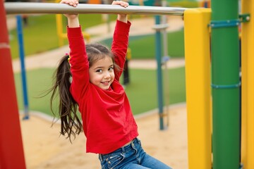 Smiling girl in red sweater hangs from monkey bars at a colorful playground with a bright background, enjoying outdoor play and fun. Ai generative