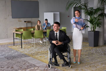 In a professional office setting, a businesswoman helping her director, who is sitting in a wheelchair and using a tablet, while their colleagues collaborate in the background.