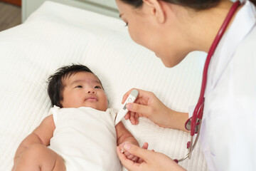 Obraz premium Caucasian female pediatrician checking temperature of Asian newborn girl using digital thermometer under arm while gently talking and smiling during healthcare checkup on white bed in clinic