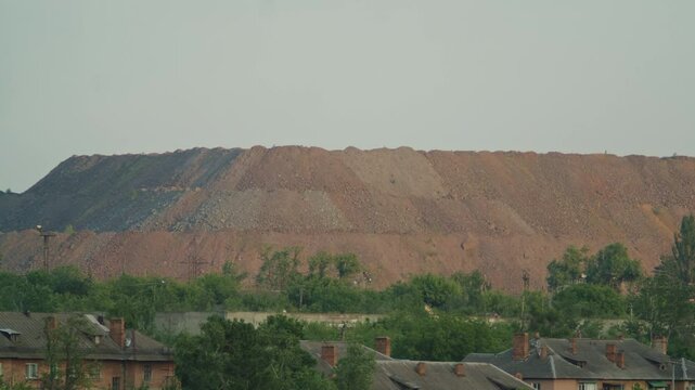 Large industrial waste heap or slag heap from mining operations near a residential area with trees and brick houses. Environmental impact and industrial landscape concept.