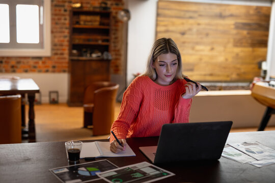 Young woman working from home, using laptop and taking notes during phone call - Powered by Adobe