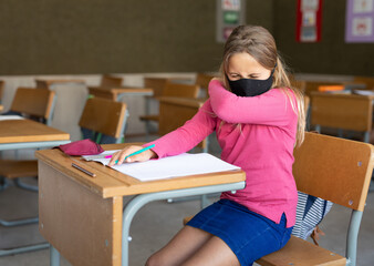 In school, young girl wearing mask sneezing into elbow at desk