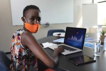 Businesswoman wearing mask using laptop in office, analyzing data on screen