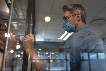 Businessman wearing mask writing on glass board in modern office environment