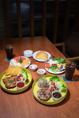 A set of pork and chicken steaks, bread and a plate of salad on a brown wooden table in a restaurant.