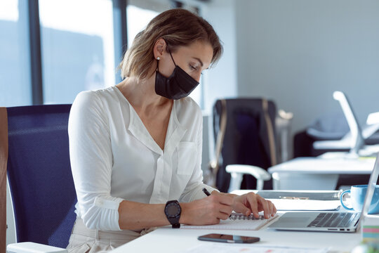 Businesswoman wearing mask writing in notebook at office desk with laptop