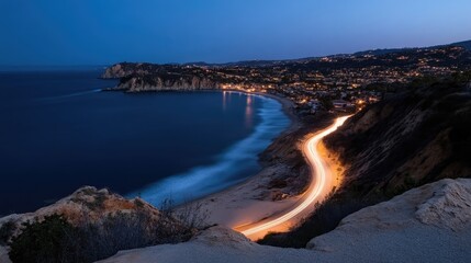 Fototapeta premium Winding coastal road with glowing car light trails curves along a sandy beach and bay next to a lit seaside town at twilight.