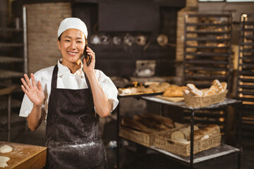Asian female baker in bakery talking on phone and smiling, surrounded by bread, copy space