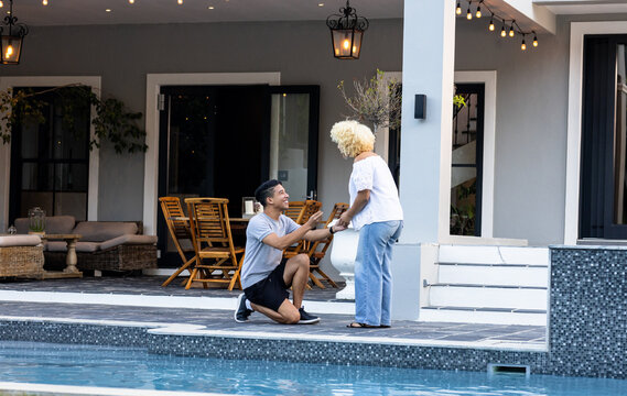 Diverse couple, Young man proposing by poolside, holding ring, woman surprised and smiling