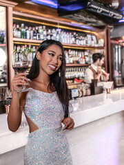 A glamorous woman in a sparkling sequin dress holding a glass of red wine at an elegant bar. She...