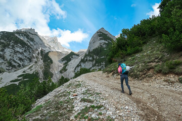 Woman hiker with backpack and helmet walks uphill on rocky trail surrounded by lush greenery,...