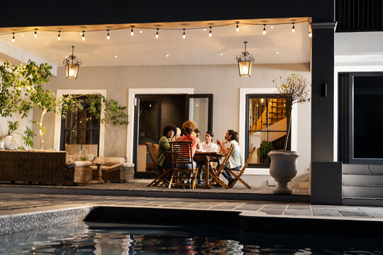 Diverse friends enjoying dinner together on patio by pool under string lights