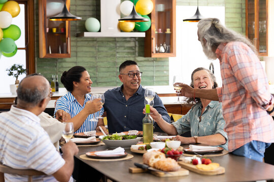 Diverse senior friends enjoying dinner party at home, laughing and toasting with wine