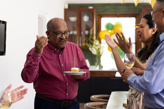 Senior man celebrating birthday with diverse friends, holding cupcake and smiling, at home