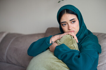 Sad muslim woman wearing hijab sitting on sofa at home hugging pillow feeling depressed